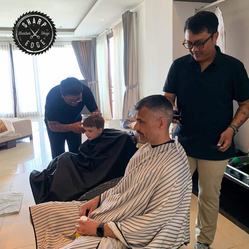 Sharp Edge Barber's VIP service, providing personalized haircuts at a client's home. A skilled barber works attentively on a young boy's haircut, while another barber perfects the hairstyle of an adult client seated nearby. The private and relaxed setup showcases Sharp Edge Barber’s commitment to high-quality, tailored grooming experiences in a comfortable setting.